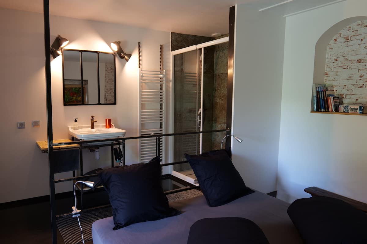 Une chambre moderne avec un grand lit, une table de chevet, un miroir, un lavabo intégré et un espace douche.