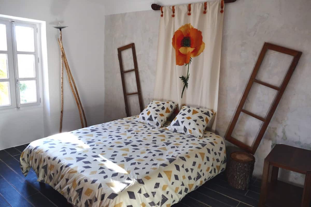 Une chambre à coucher avec un lit, une fenêtre, une lampe et un rideau orné de motifs floraux.