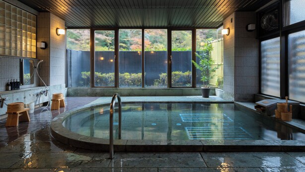 Uma onsen japonesa tradicional com um deck de madeira, uma banheira de pedra e vista para o verde ao redor.