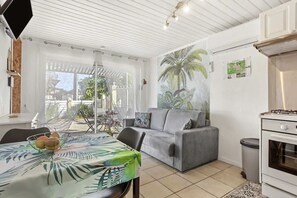 A modern kitchen with a stove, a dining table with a fruit bowl, a grey sofa, and a large window with a view of the outdoors. - Homerez - Holiday Apartment in Vidauban (Vidauban)