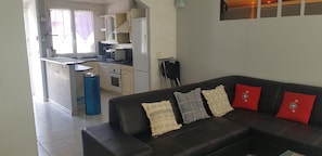 A living room with a black leather sofa, a kitchen with a stove and refrigerator, and a window with curtains.