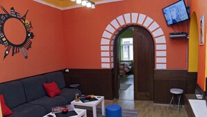 A room with a grey sofa, red cushions, a white table, a blue stool, and a TV on the wall. - Traveler's Hostel & Apartments (Belgrade)
