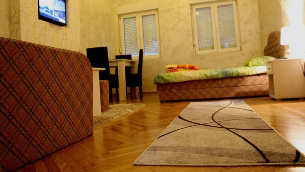 A room with a patterned sofa, a small desk with a chair, a bed with a green blanket, and a television mounted on the wall. - Traveler's Hostel & Apartments (Belgrade)