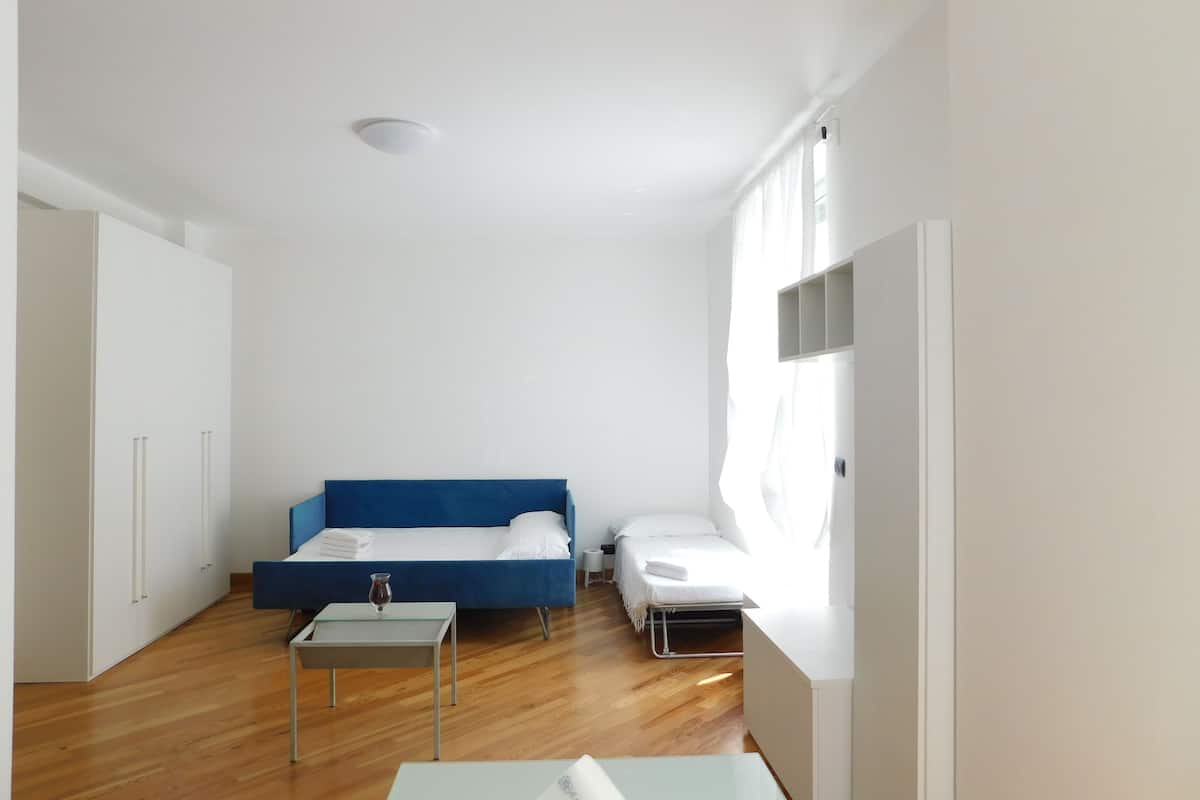 Une petite pièce lumineuse avec un lit bleu, une table de chevet et une petite fenêtre.