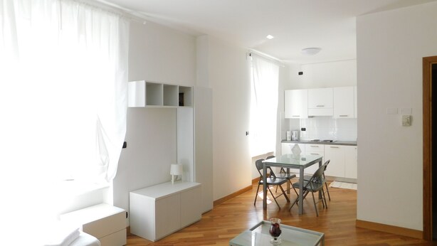 Ein modernes, helles Wohnzimmer mit einem Glastisch, einem Essbereich mit Tisch und Stühlen sowie einer Küche im Hintergrund.