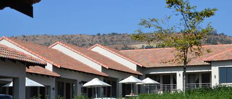 A row of hotel rooms with red-tiled roofs, outdoor umbrellas, and a view of a hillside.