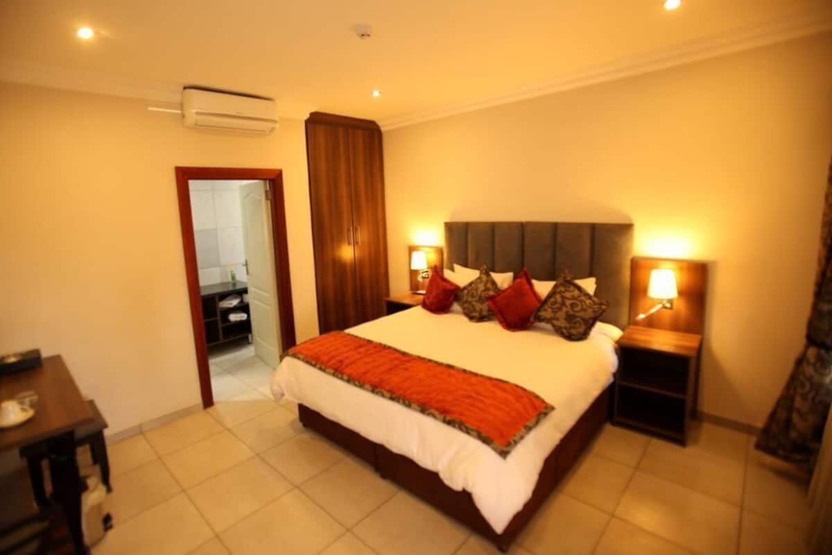 Habitación de hotel con una cama grande, dos mesitas de noche con lámparas, un equipo de aire acondicionado y vista al baño a través de una puerta abierta.