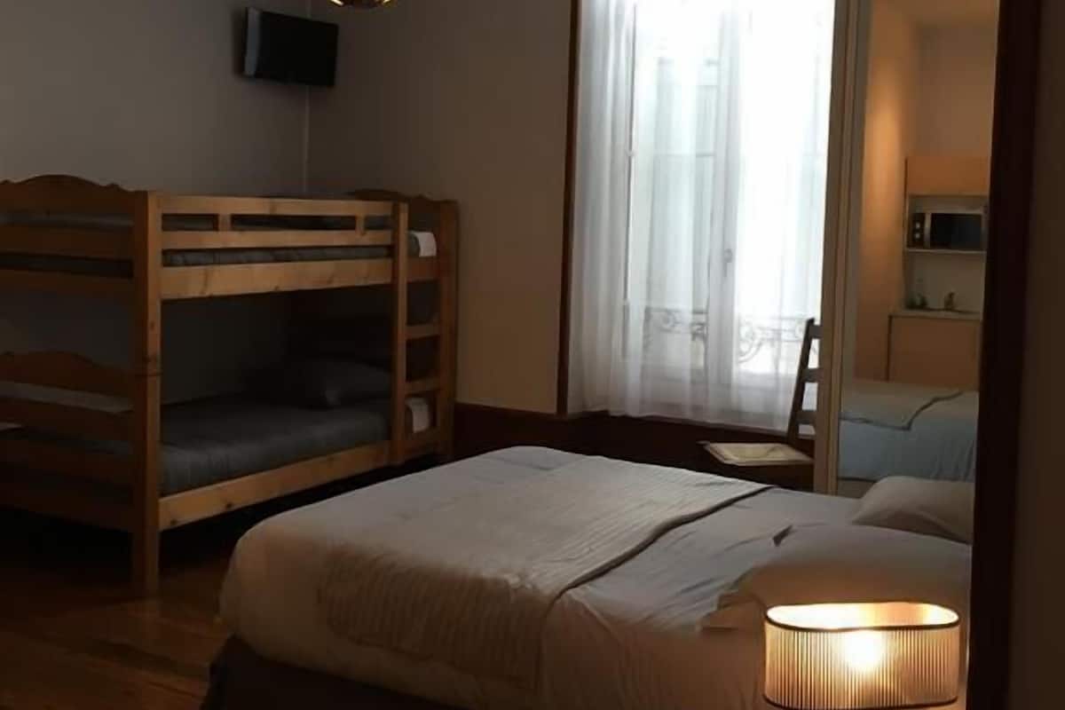 A hotel room with a bunk bed, a single bed, a bedside table with a lamp, a TV on the wall, and a window with curtains.