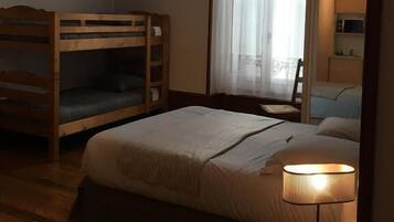 A hotel room with a bunk bed, a single bed, a bedside table with a lamp, a TV on the wall, and a window with curtains.