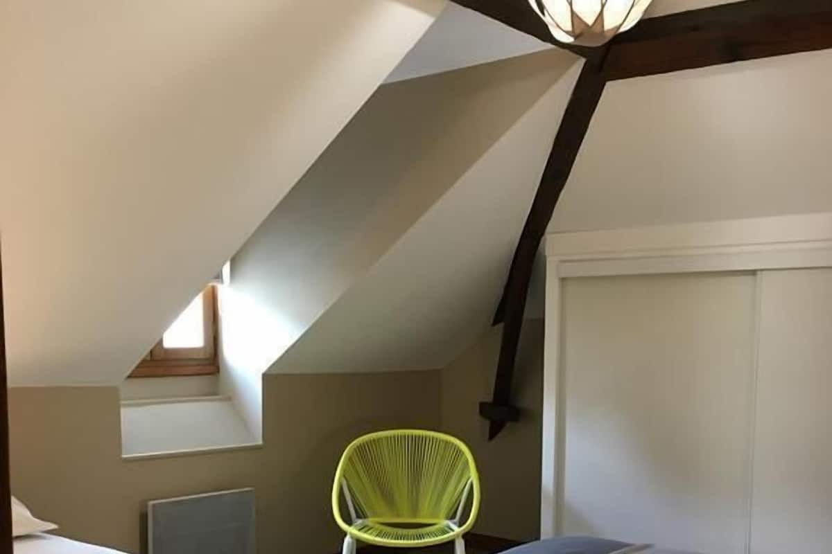 A small, cozy attic bedroom with two beds, a bright green chair, and a sloped ceiling with exposed beams.