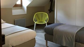 Une petite chambre mansardée, chaleureuse et accueillante, avec deux lits, un fauteuil vert vif et un plafond en pente avec des poutres apparentes.