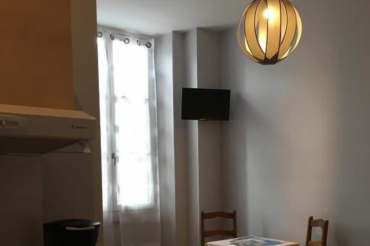 A compact hotel room with a bed, a small table with a chair, a coffee maker, a TV mounted on the wall, and a window with curtains.