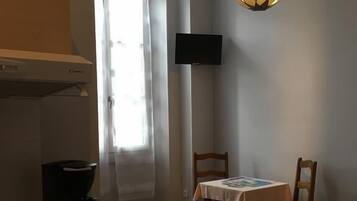 Habitación de hotel pequeña con cama, una mesita con una silla, cafetera, televisor montado en la pared y ventana con cortinas.