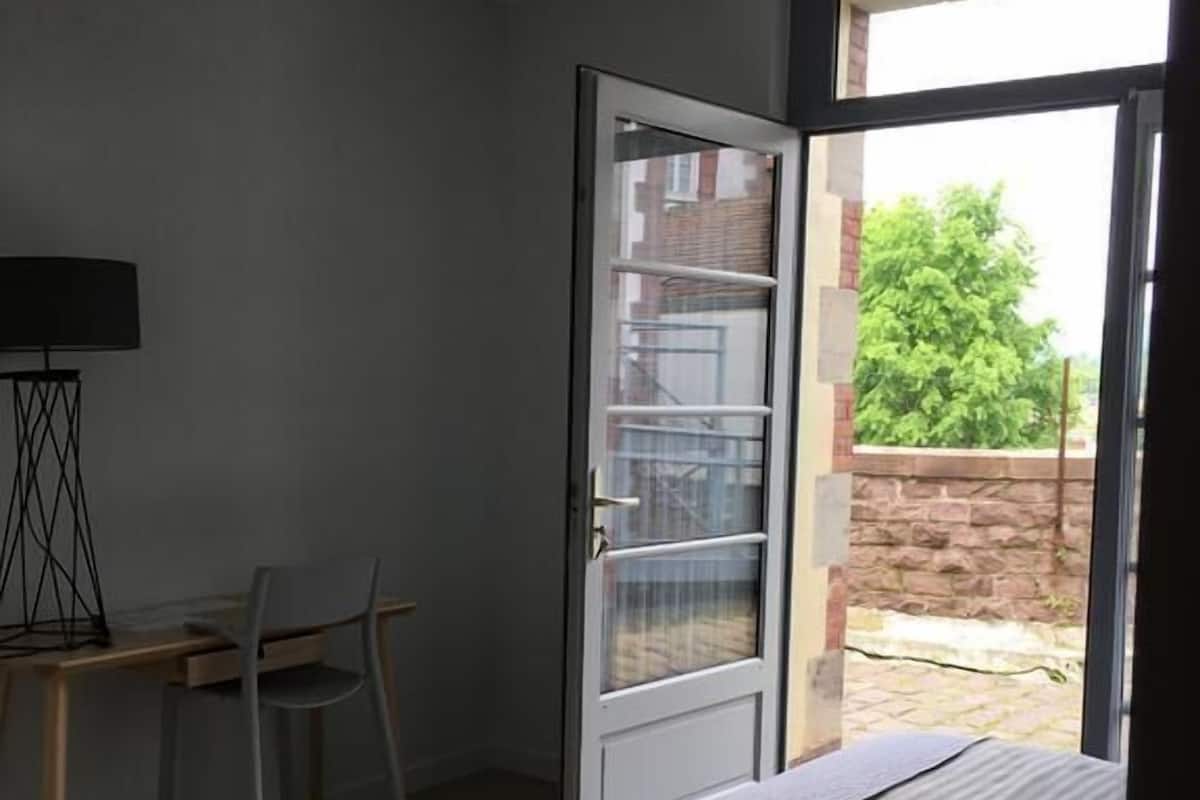 A bedroom with a bed, a desk with a chair, and a window opening to an outdoor area.