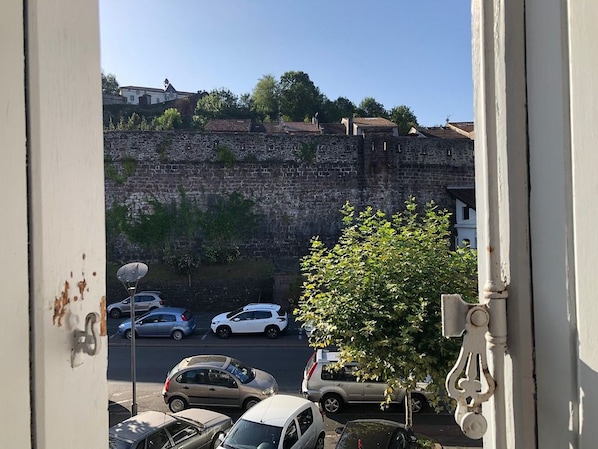 Vista a través de una ventana que muestra una calle con coches aparcados, un muro de piedra y edificios al fondo.
