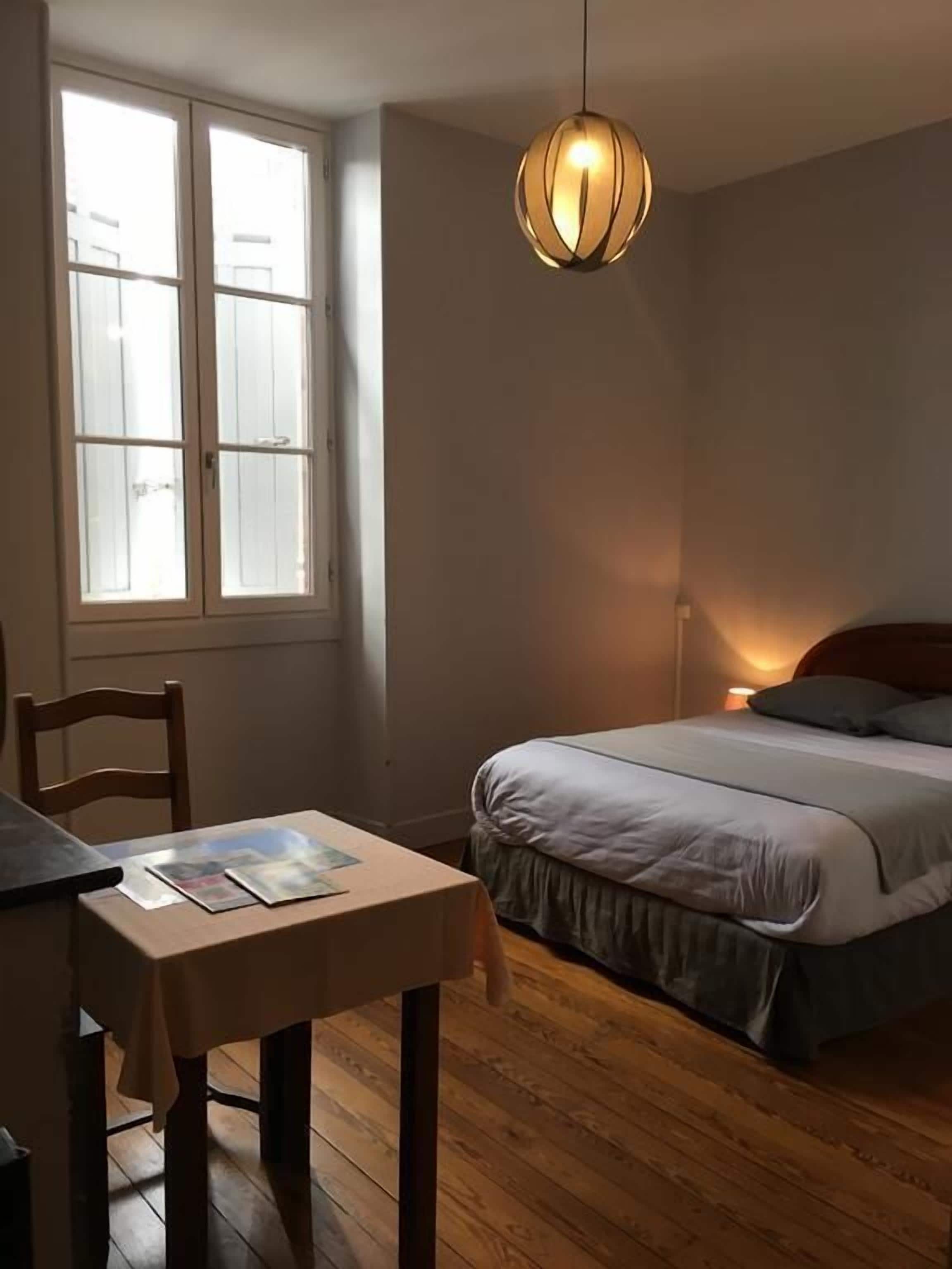 A bedroom with a bed, a table with a chair, a window, and a hanging light fixture.