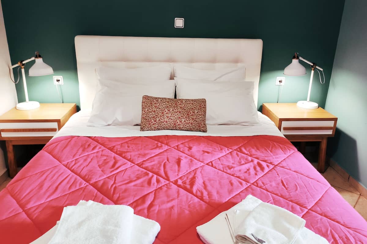 Cama com colcha rosa, almofadas brancas e uma almofada de pelo de leopardo. Há duas mesas de cabeceira com abajures.