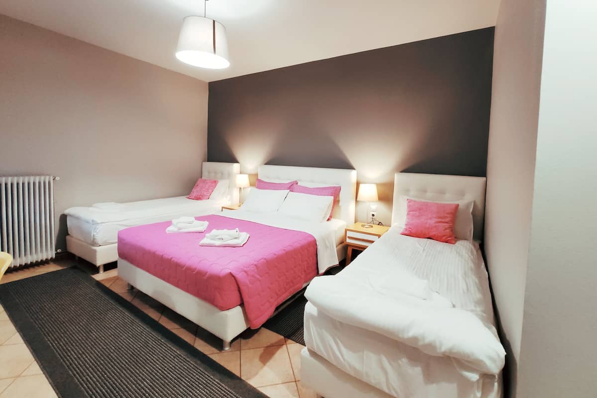 Quarto de hotel com duas camas, um radiador, uma mesa de cabeceira com abajur e uma janela grande.