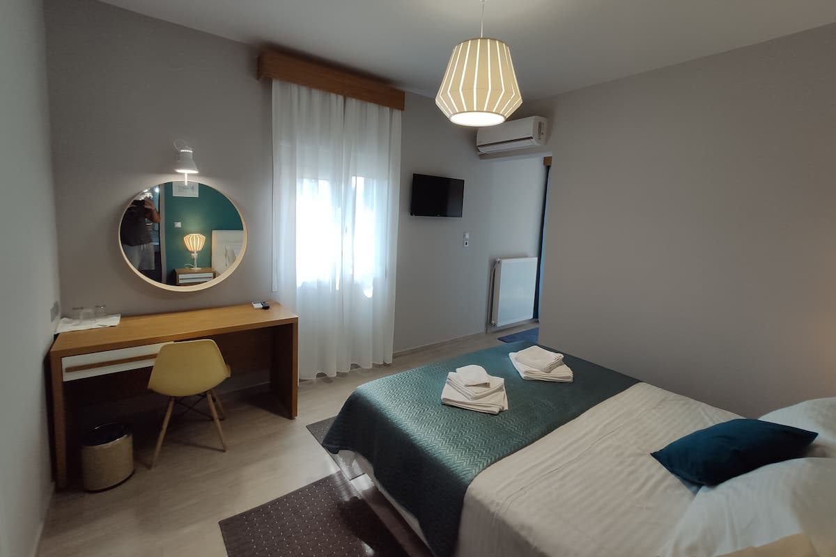Quarto de hotel com cama, escrivaninha, cadeira, espelho redondo, luminária pendente e televisão.