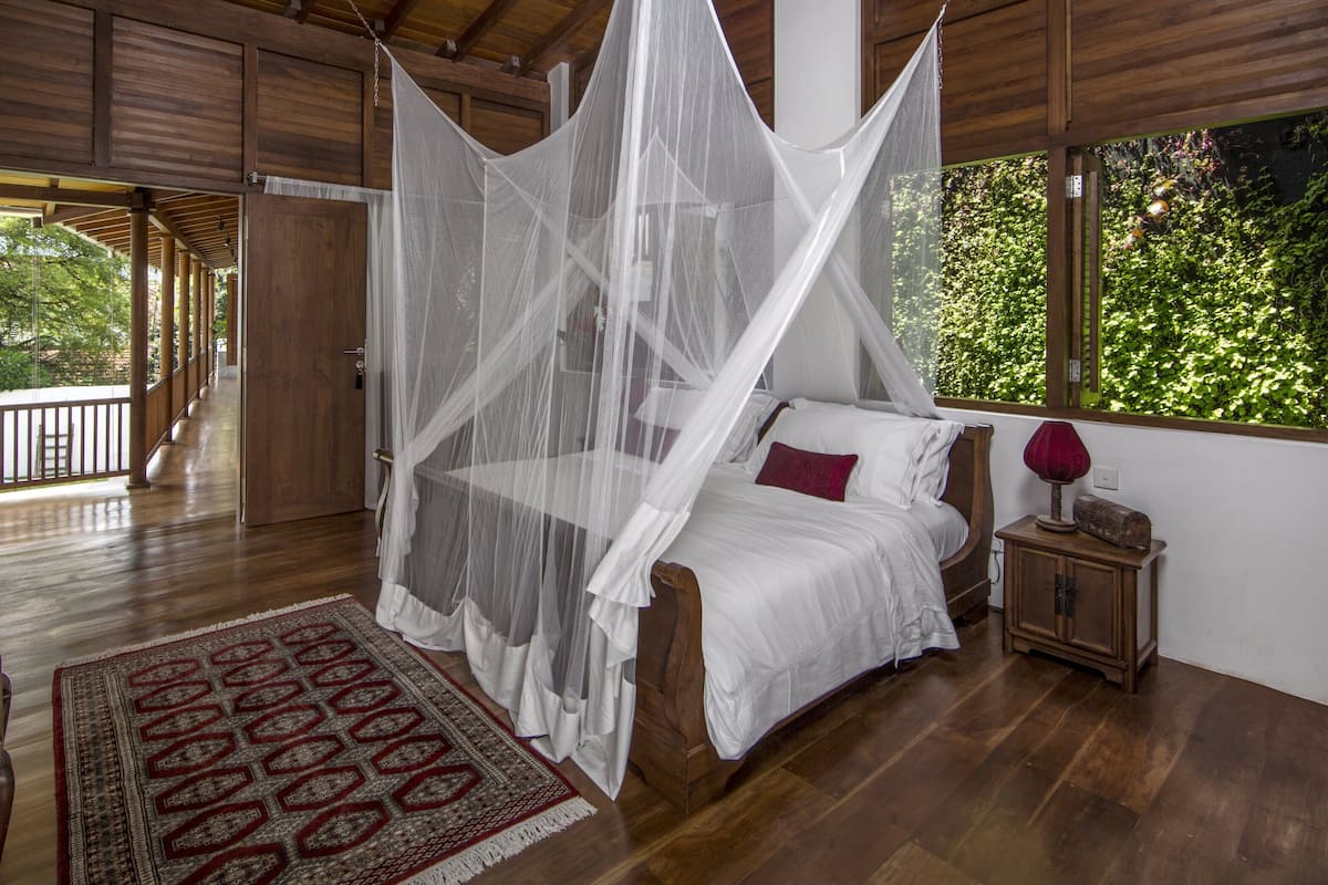 A bedroom with a four-poster bed, a wooden floor, a red bench, a patterned rug, and a view of greenery through the window.