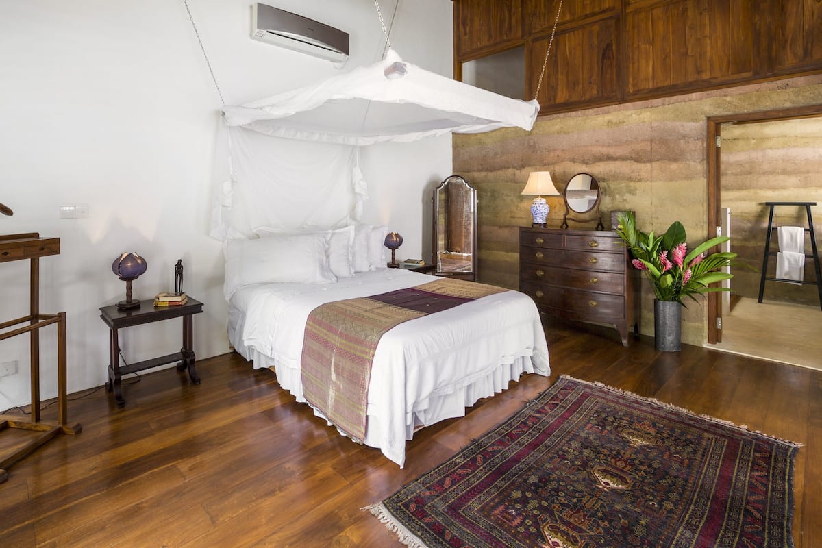 A bedroom with a bed, bedside table, dresser, mirror, hanging clothes rack, and a large window.