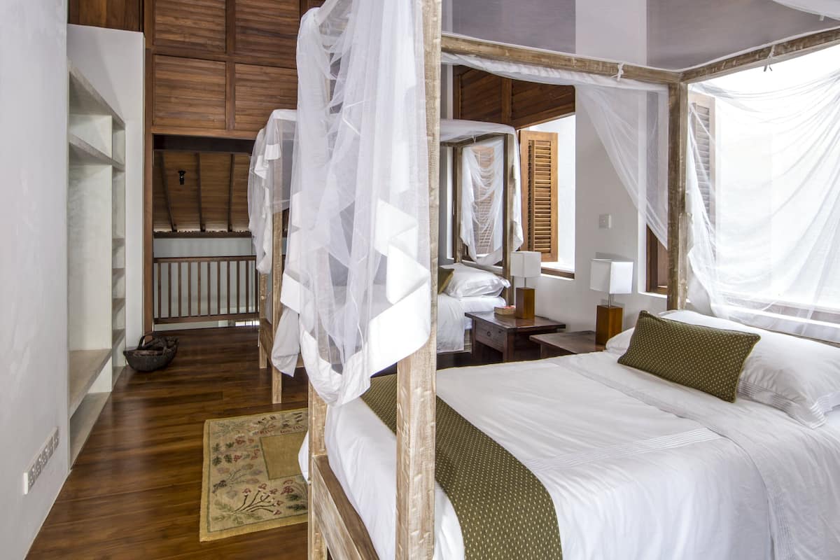 A bedroom with a wooden bed frame, white bedding, and a green pillow. There is a nightstand with a lamp, a window with curtains, and a wooden floor.