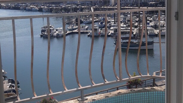 Une vue depuis un balcon donnant sur une marina où sont amarrés des bateaux, avec une piscine carrelée et des bâtiments en arrière-plan.