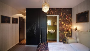 A hotel room with a bed, a chandelier, a bathroom with a sink, and a painting of apples on the wall. - Das kleine Schwarze (Hamburg)
