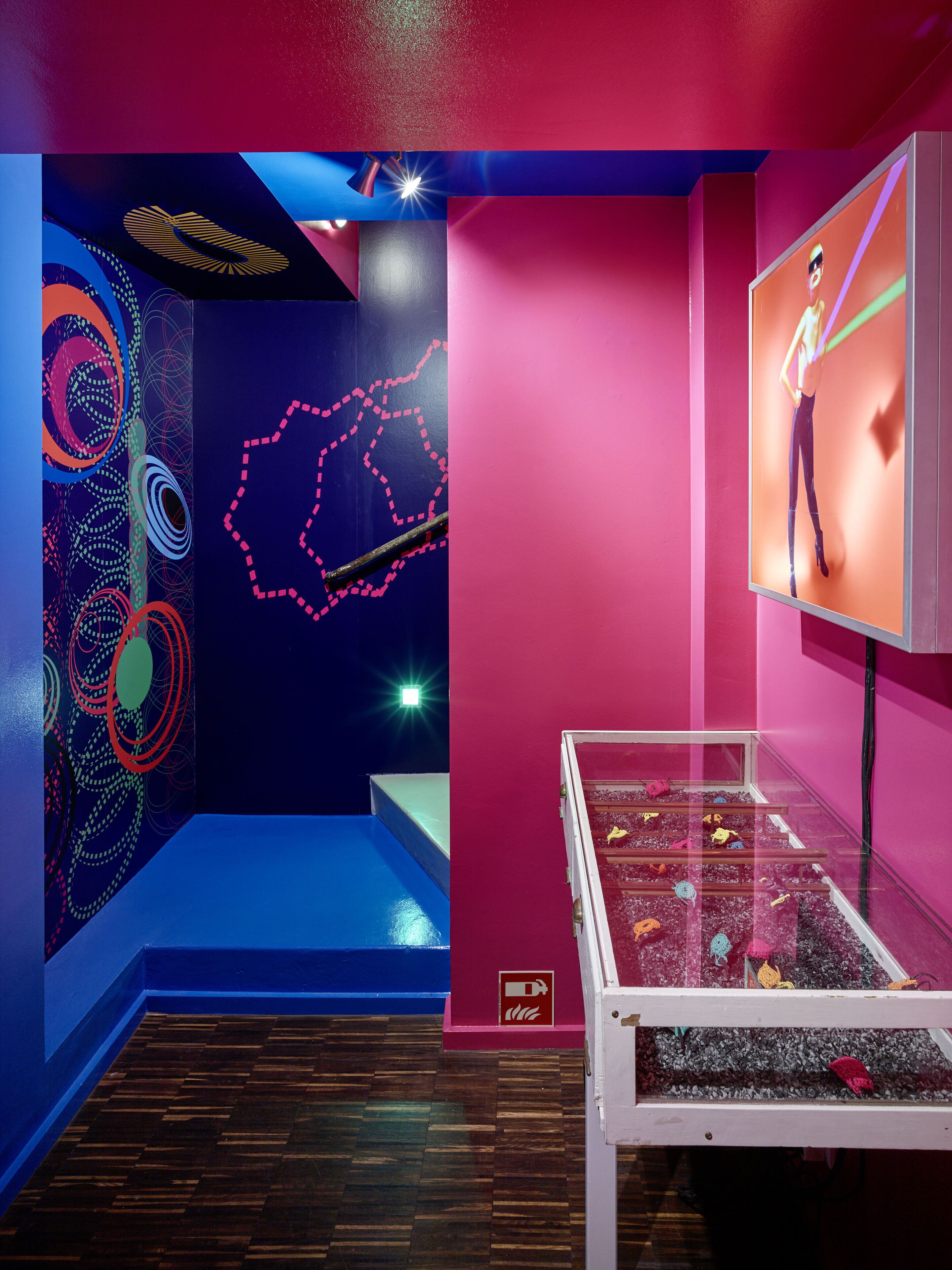Una galería de arte moderno con diseños abstractos en las paredes, una pantalla digital y una vitrina de cristal con objetos coloridos.