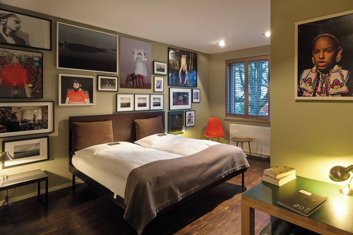 Un dormitorio con cama, escritorio y silla. Hay cuadros en la pared y una ventana con persianas.