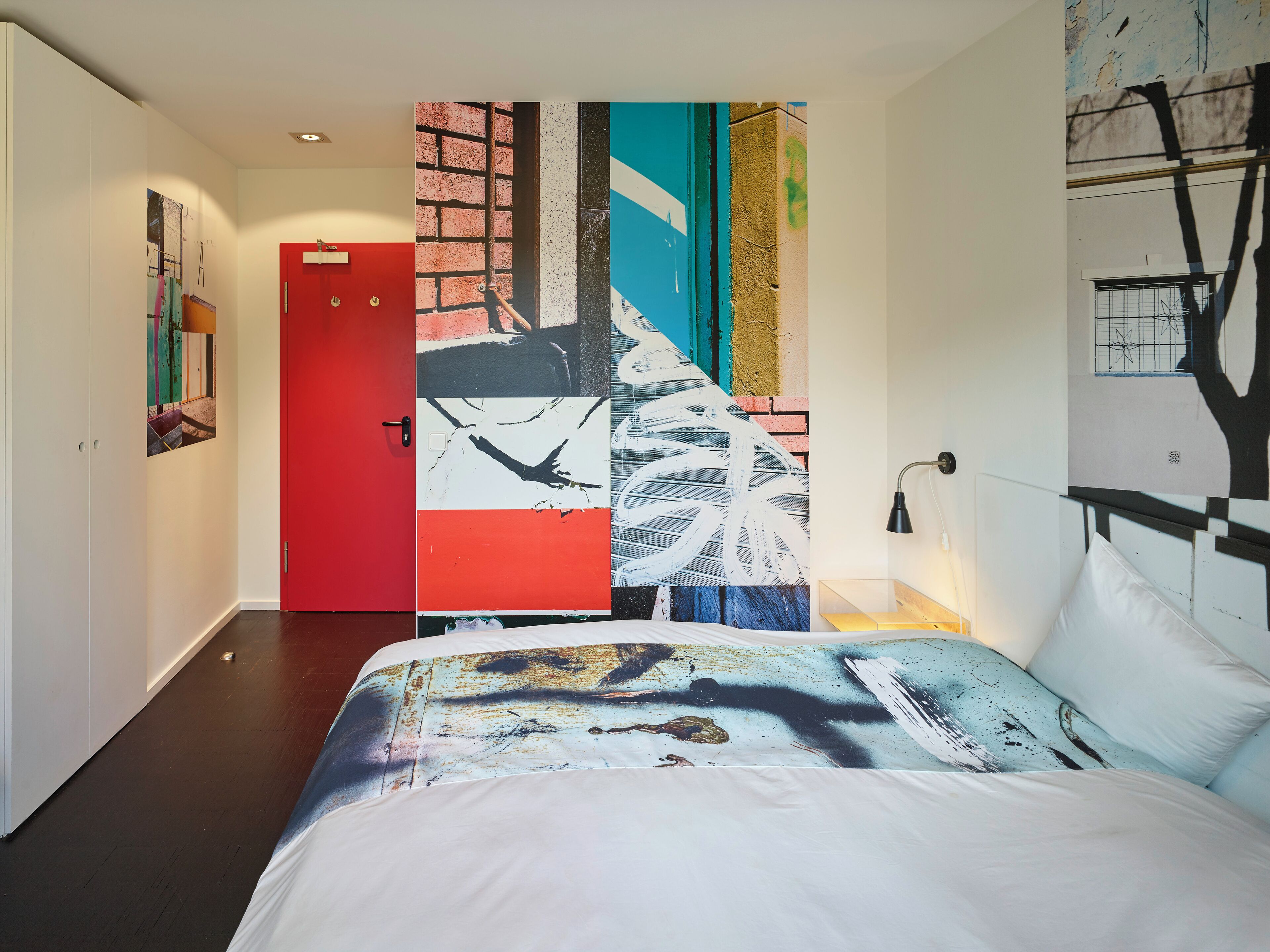 A modern hotel room with a large bed, a red door, and a wall with abstract art.