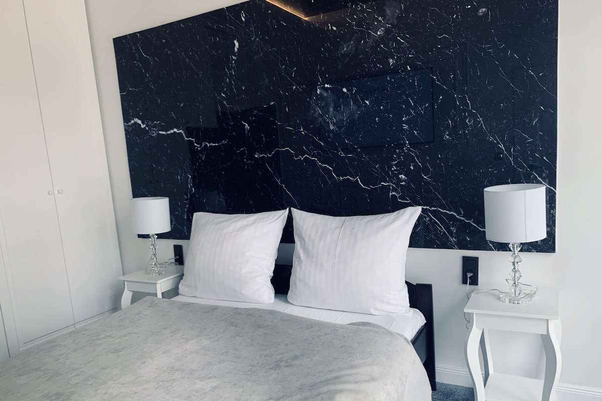 Une chambre à coucher moderne avec un grand lit, une literie blanche, deux tables de chevet avec des lampes et une tête de lit en marbre.