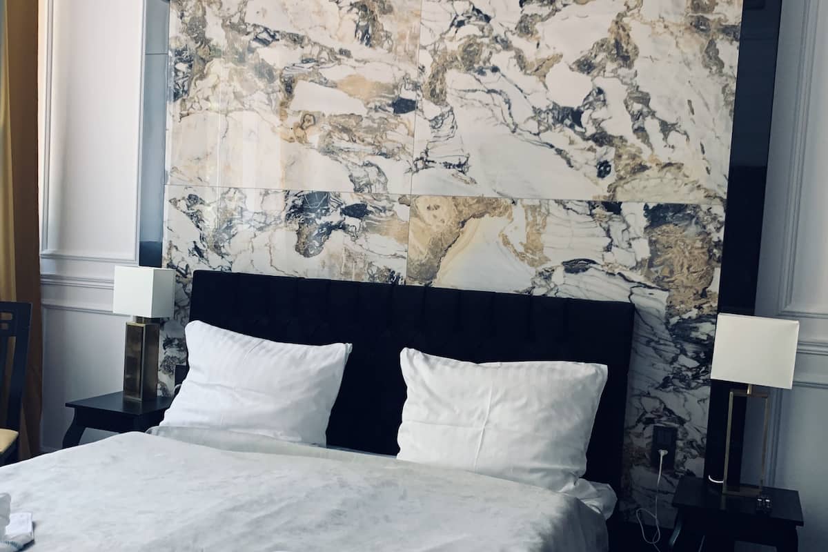 Un grand lit avec du linge de lit blanc et des oreillers, une tête de lit à motifs de marbre et deux tables de chevet avec des lampes.