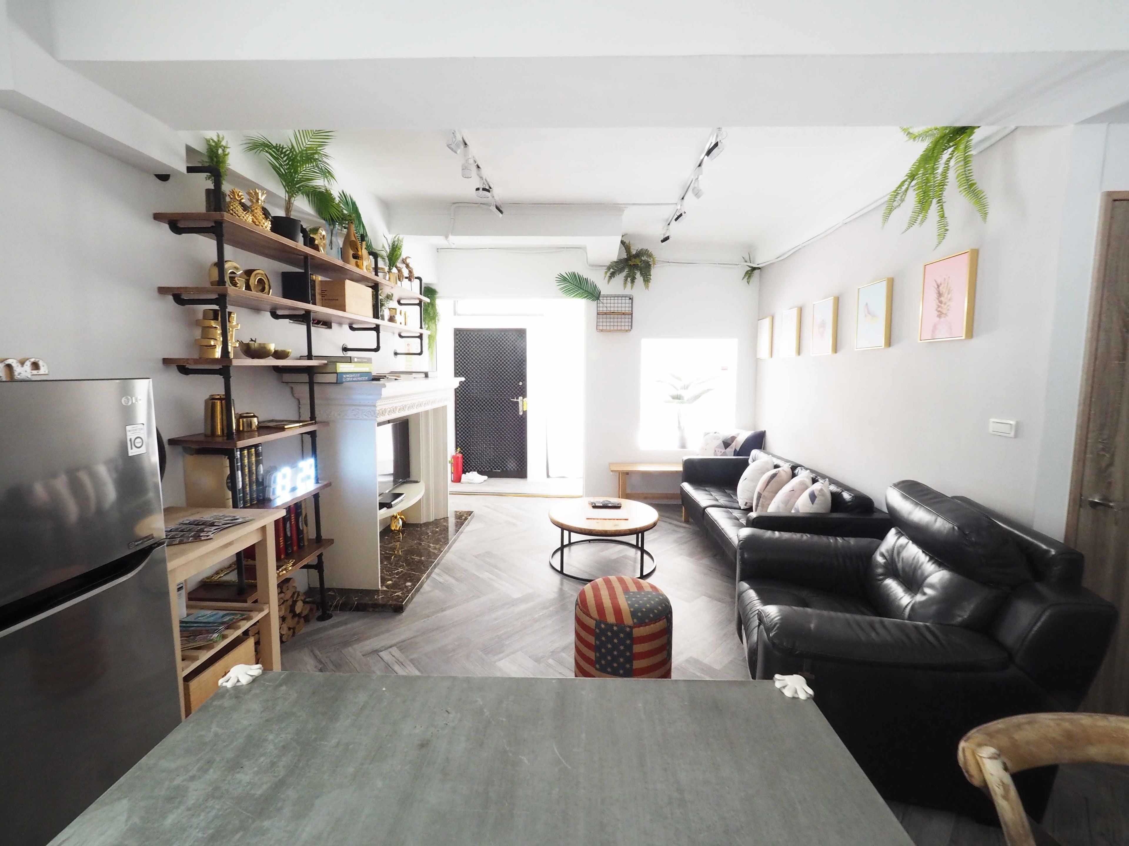 A modern living room with a black leather sofa, a wooden coffee table, and a dining table. There are shelves with decorative items and a small American flag-patterned ottoman.