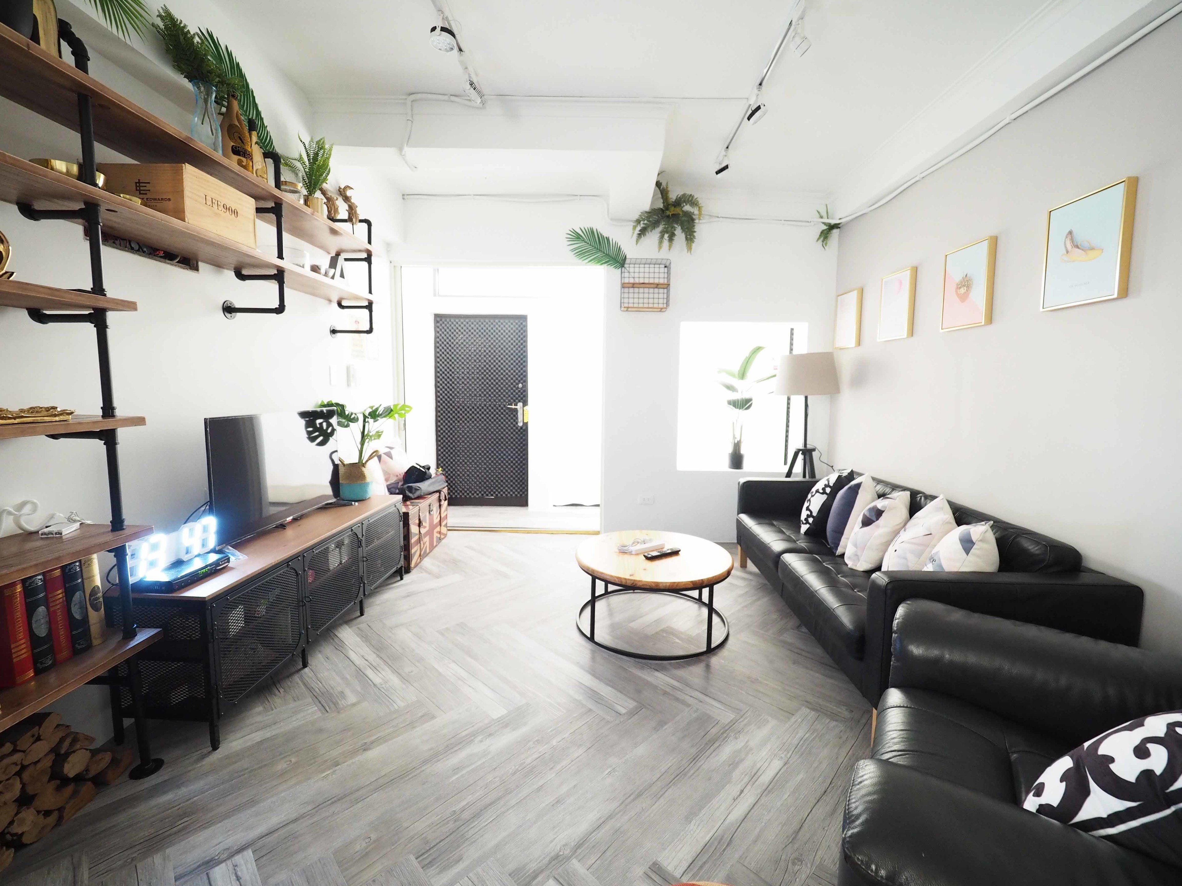 A modern living room with a black leather sofa, a wooden coffee table, and a TV on a stand. There are shelves with decorative items and plants, a black door, and a window with a view.