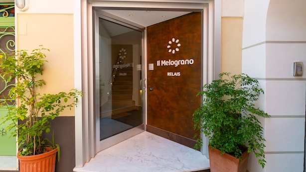 A hotel entrance with a glass door, a wooden panel with the hotel name, and two potted plants. - Il Melograno Relais (Vico Equense)