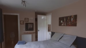 A bedroom with a large bed, a bedside table, a wall-mounted TV, and a door leading to another room. - Grandwood House (Chichester)