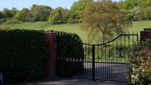 A gated entrance with a brick wall and metal gate, leading to a green field with trees. - Grandwood House (Chichester)