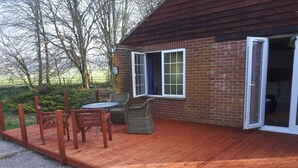 A wooden deck with a table and chairs, a brick building with a sliding glass door, and a view of trees and a field. - Grandwood House (Chichester)