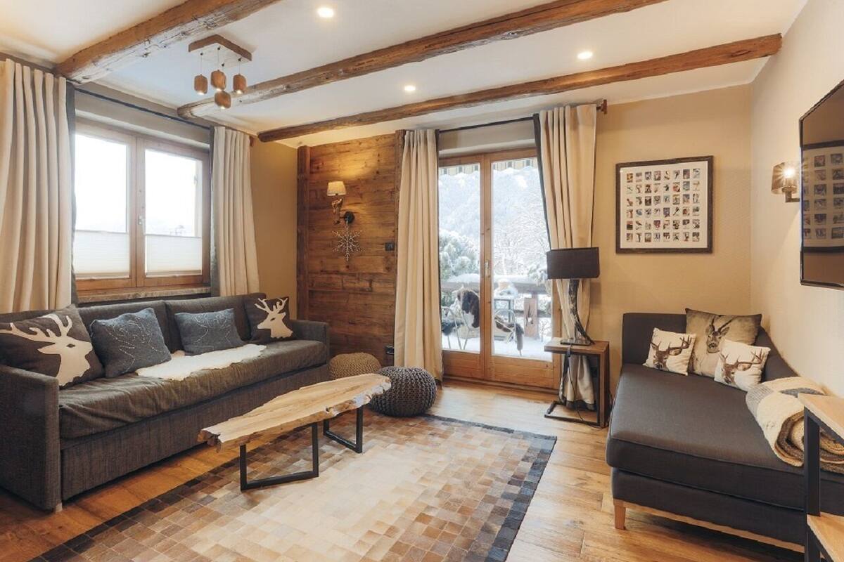 A cozy living room with a sofa, a bench, and a bed. There is a fireplace, a window, and a wall with framed pictures.