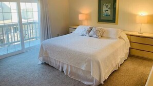 A hotel room with a large bed, two bedside tables, a ceiling fan, a balcony with a view, and a framed picture on the wall. - LITTLE is only 1 block walk to beach access - 4 BR - sleeps 8 (Duck)