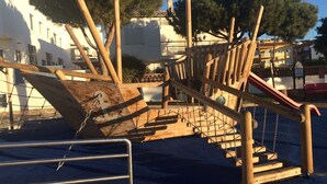 Une structure de jeux en bois, dotée d’un toboggan et d’éléments d’escalade, entourée d’arbres et de bâtiments.