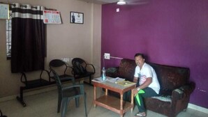 A room with a purple wall, a ceiling fan, a sofa, a table, and two chairs. - iROOMZ Pranathi Comforts (Ballari)