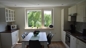 Een keuken met een eettafel, stoelen, een magnetron, kastjes en een raam met uitzicht op bomen.