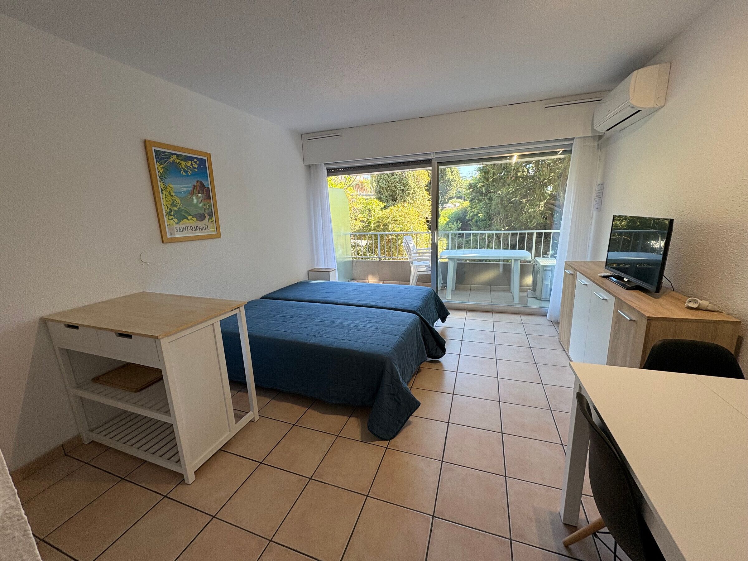 A small, clean room with two beds, a desk, a TV, and a balcony with a view of trees.