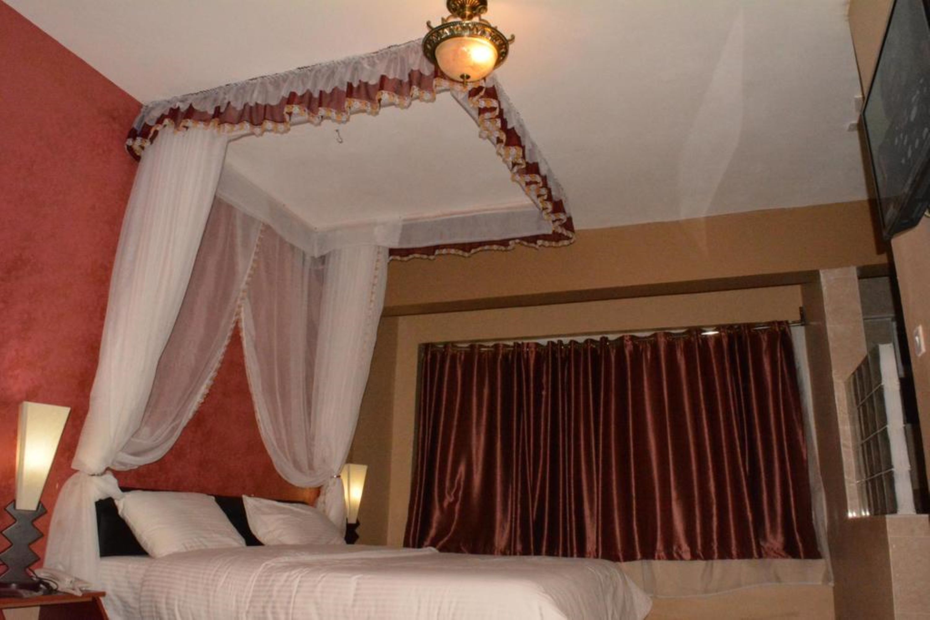 A bedroom with a canopy bed, a large window with curtains, and a wall-mounted TV.