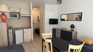 Cocina compacta con microondas, un refrigerador pequeño y un lavamanos. Hay una zona de comedor con mesa y sillas, y un sofá con cojines. Un televisor de pantalla plana está montado en la pared.