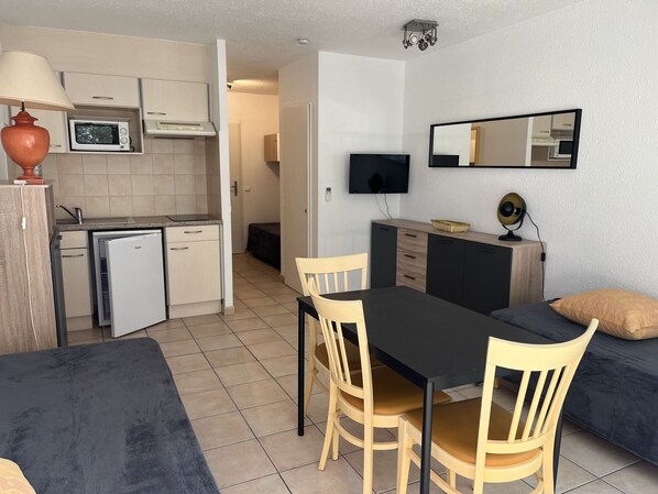 A compact kitchen with a microwave, a mini-fridge, and a sink. There is a dining area with a table and chairs, and a sofa with cushions. A flat-screen TV is mounted on the wall.