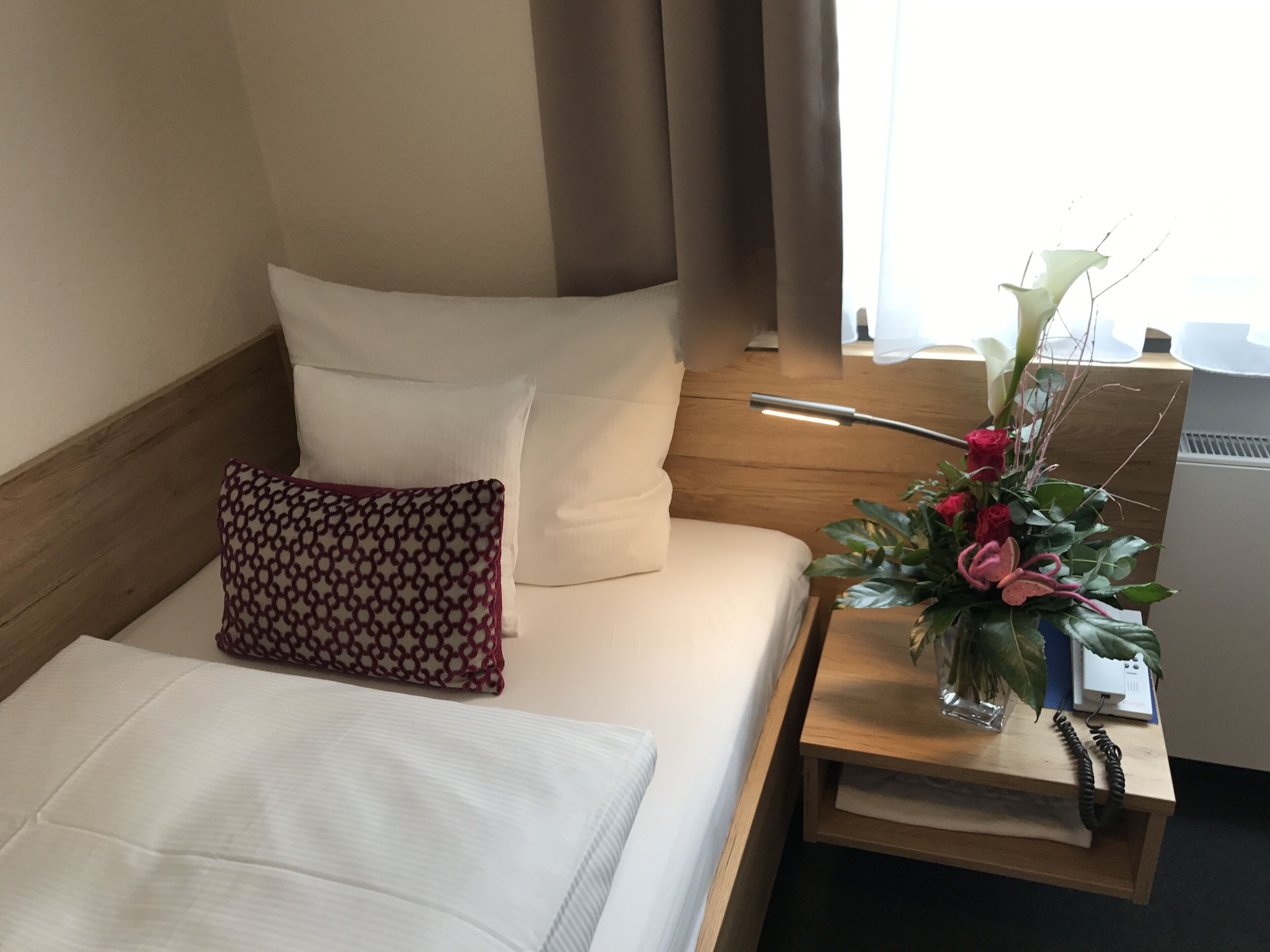 A hotel room with a neatly made bed, a bedside table with a lamp, a vase with flowers, and a power outlet on the nightstand.