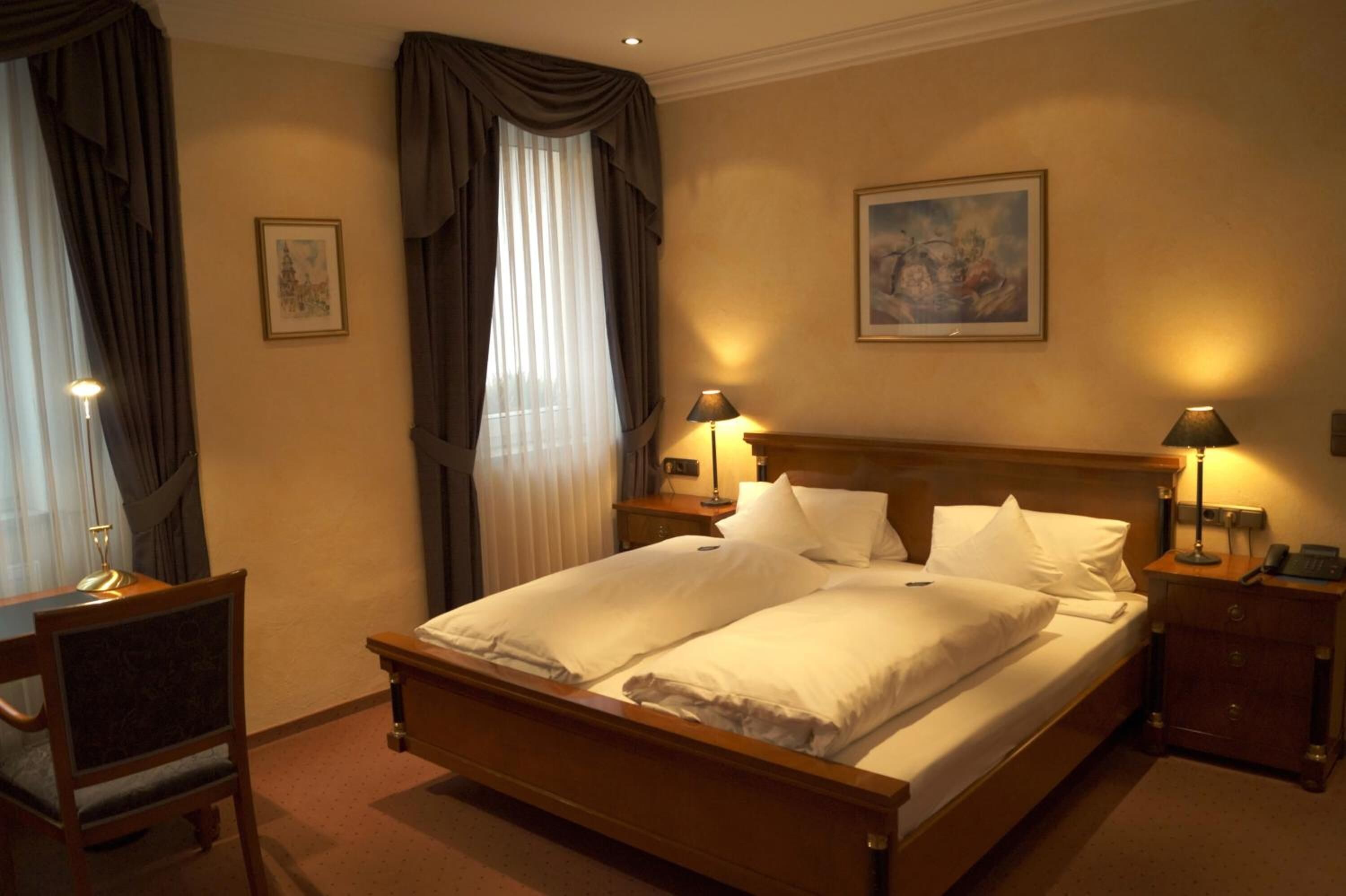 A hotel room with a double bed, two bedside tables with lamps, a desk with a chair, a window with curtains, and a painting on the wall.